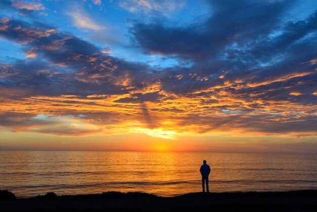 Sylwetka osoby stojącej na plaży pod zachmurzonym niebem podczas zapierającego dech w piersiach zachodu słońca