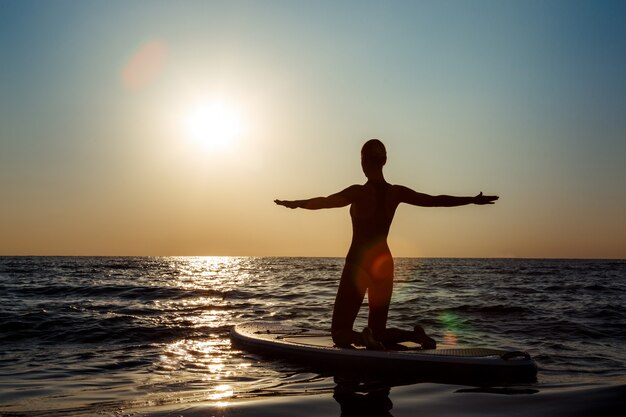 Sylwetka pięknej kobiety ćwiczy joga na surfboard przy wschodem słońca.