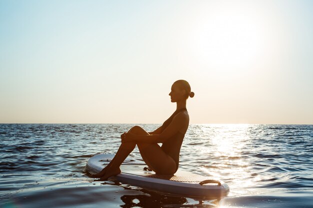 Sylwetka pięknej kobiety ćwiczy joga na surfboard przy wschodem słońca.