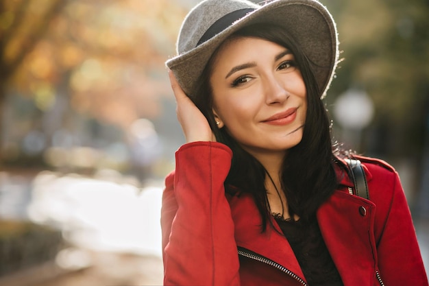 Szczegół portret czarnowłosa kobieta z makijażem nago pozowanie z przyjemnością na tle lasu rozmycie. Uśmiechnięta pani w kapeluszu i czerwonym płaszczu wyrażająca prawdziwe emocje.