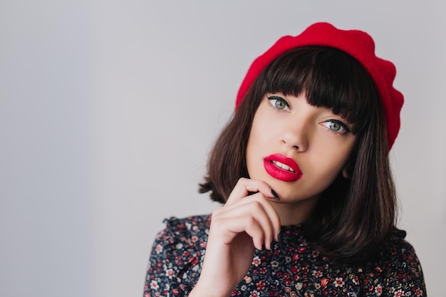 Szczegół portret elegancka brunetka dziewczyna z krótkimi włosami na sobie czerwony beret i dotykając jej podbródka. Piękna młoda ciemnowłosa kobieta w stroju vintage pozuje medytująco na białym tle na jasnym tle