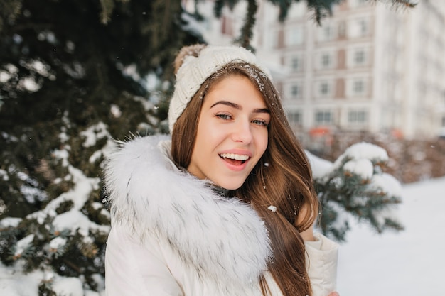 Szczegół portret kobiety o niebieskich oczach ze śniegiem we włosach, ciesząc się szczęśliwy czas zimowy. Plenerowe zdjęcie zmysłowej blondynki ze szczerym uśmiechem stojącej na ulicy obok zielonego świerku.