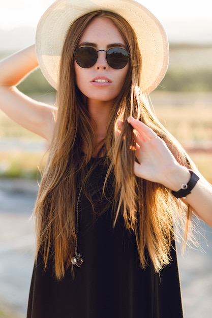 Szczegół portret pięknej młodej dziewczyny z długimi ciemnymi włosami na sobie słomkowy kapelusz, ciemne okulary przeciwsłoneczne. Bawi się włosami w ciepłych promieniach zachodzącego słońca