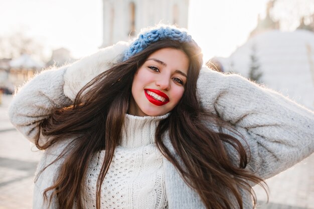 Szczegół portret wesoła dziewczyna z długimi czarnymi włosami pozowanie w zimowy poranek na miasto rozmycie. Brunetka dama w niebieskim berecie podczas sesji zdjęciowej w zimny dzień.