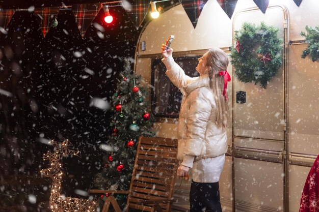 Szczera młoda kobieta robi selfie trzymając w rękach smartfon i ciesz się szczęśliwego nowego roku i świąt bożego narodzenia ferie zimowe w wigilię noc na kempingu podróż przyczepa dom na zewnątrz. spadający śnieg