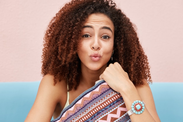 Szczere ujęcie śmiesznej Afro American młodej kobiety robi grymas, zaokrągla usta, ma krzaczaste kręcone włosy