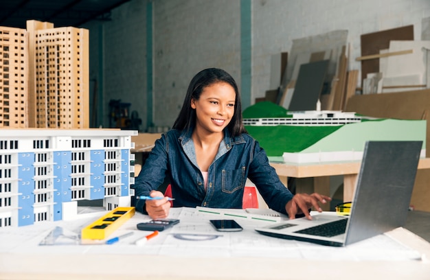 Szczęśliwa afroamerykańska dama z laptopem i modelem budynek przy stołem
