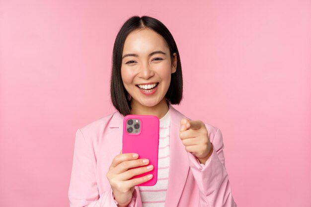 Szczęśliwa azjatycka bizneswoman śmiejąca się wskazującym na ciebie palcem i nagrywająca wideo robiąca zdjęcia na smartfonie za pomocą różowego tła telefonu komórkowego