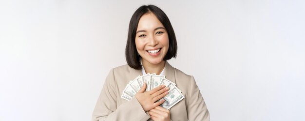 Szczęśliwa azjatycka bizneswoman trzymająca gotówkę przytulająca dolary pieniądze i uśmiechnięta stojąca na białym tle w garniturze