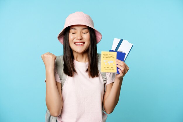 Szczęśliwa Azjatycka dziewczyna triumfuje, pokazując świadectwo szczepień, paszport zdrowia i bilety lotnicze, świętując podróż, turysta czuje radość, stojąc na niebieskim tle
