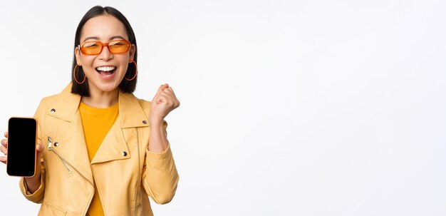 Szczęśliwa azjatycka dziewczyna w okularach przeciwsłonecznych pokazująca interfejs smartfona z ekranem telefonu komórkowego śmiejąca się i uśmiechnięta świętująca stojąca na białym tle