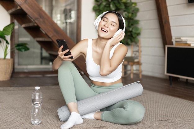 Szczęśliwa azjatycka dziewczyna w stroju fitness siedzi w domu na podłodze słuchając muzyki i ćwicząc, trzymając sm...