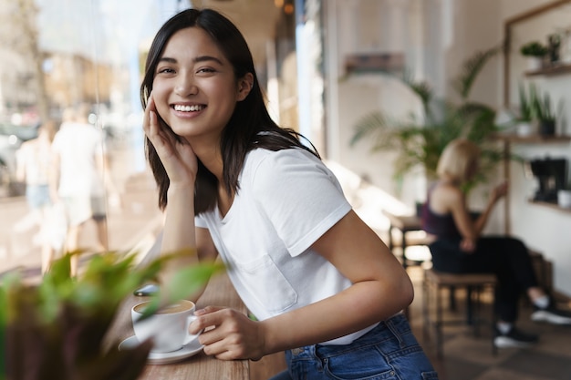 Szczęśliwa azjatycka kobieta siedzi w restauracji w pobliżu okna i uśmiecha się do kamery.