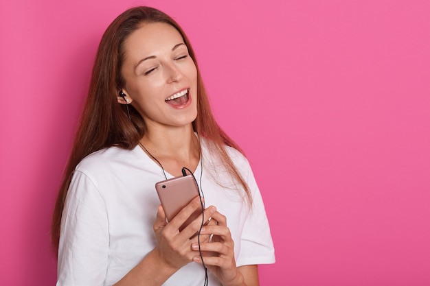 szczęśliwa beztroska młoda kobieta tańczy i słucha muzyki ze smartfona na różowym tle, pani ubrana w białą koszulę