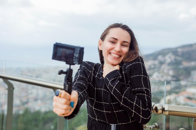 Szczęśliwa blogerka robi selfie swoim mini aparatem, trzymając rękę na klatce piersiowej na tle widoku miasta