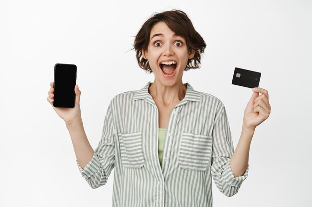 Szczęśliwa brunetka krzyczy z radości, pokazując ekran telefonu komórkowego i karty kredytowej, białe tło.