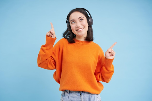 Bezpłatne zdjęcie szczęśliwa chinka w słuchawkach słucha muzyki, lubi ulubioną piosenkę na swojej liście odtwarzania, stoi nad bl