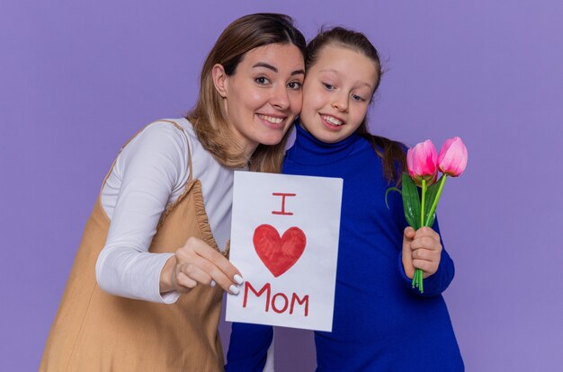 Szczęśliwa córka daje kartkę z życzeniami i kwiaty tulipanów dla niej zaskoczona i uśmiechnięta matka obchodzi dzień matki stojącej nad fioletową ścianą
