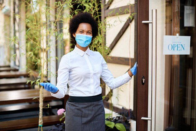 Szczęśliwa czarna kelnerka z maską ochronną ponownie otwiera kawiarnię po epidemii COVID19