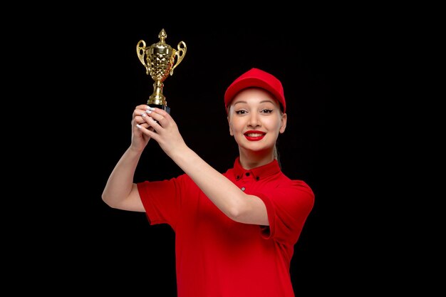 Szczęśliwa dziewczyna w czerwonej koszuli pokazująca złote trofeum w czerwonej czapce, ubrana w koszulę i jasną szminkę