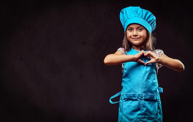 Szczęśliwa dziewczynka ubrana w mundur niebieski kucharz pokazuje gest miłości. Na białym tle na ciemnym tle z teksturą.