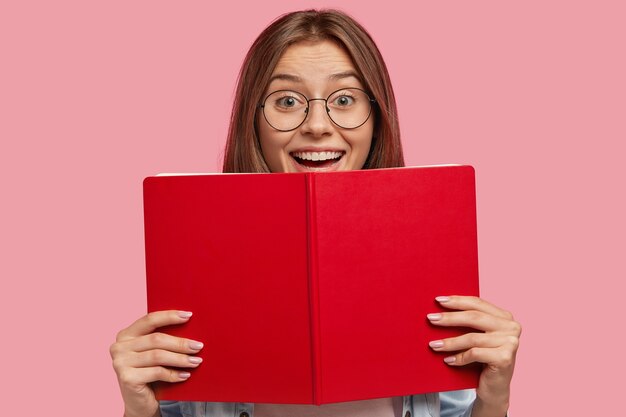 Szczęśliwa europejska studentka w okularach, ma pozytywny wyraz twarzy, trzyma czerwoną książkę, raduje się pomyślnie zdanym egzaminem na uniwersytecie, odizolowana na różowej ścianie. Ludzie, uczenie się, czytanie