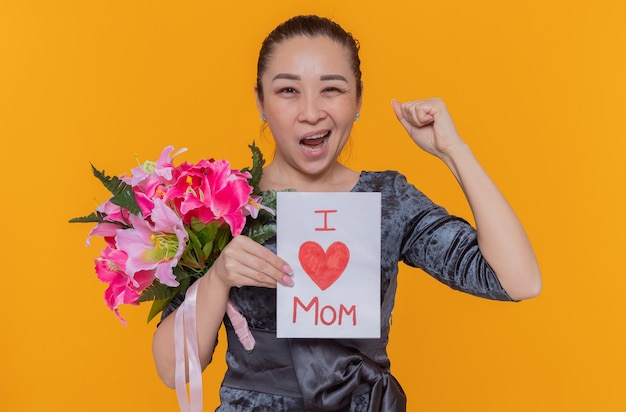 Bezpłatne zdjęcie szczęśliwa i podekscytowana azjatycka kobieta trzymająca kartkę z życzeniami i bukiet kwiatów z okazji międzynarodowego dnia kobiet zaciskająca pięść stojącą nad pomarańczową ścianą