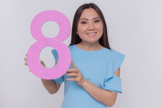Szczęśliwa i pozytywna azjatycka kobieta trzymająca numer osiem z kartonu, patrząc na przód, uśmiechnięta radośnie świętująca międzynarodowy dzień kobiet stojąca na białej ścianie