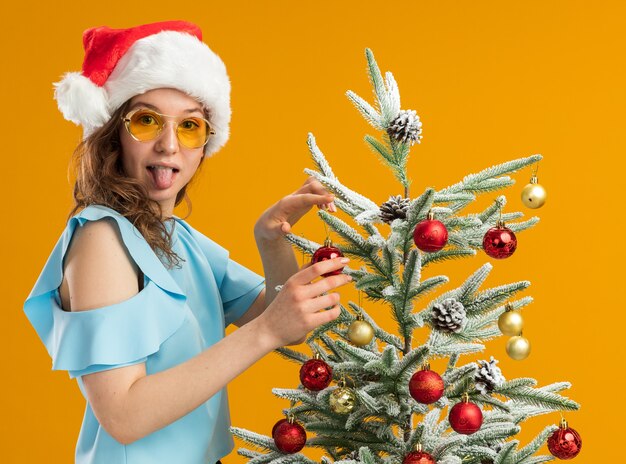 szczęśliwa i pozytywna młoda kobieta w niebieskim topie i czapce Mikołaja w żółtych okularach dekorująca choinkę wystający język patrząc w kamerę stojąca na pomarańczowym tle