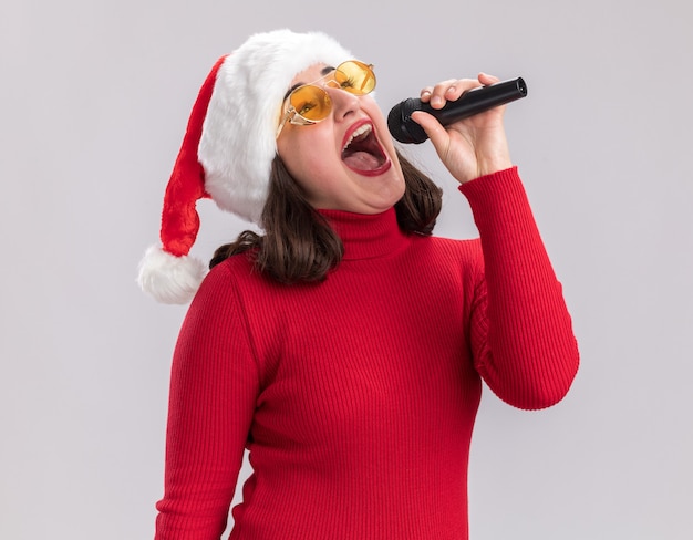 Szczęśliwa i wesoła młoda dziewczyna w czerwonym swetrze i kapeluszu santa w okularach trzymając mikrofon śpiewa piosenkę stojącą na białej ścianie