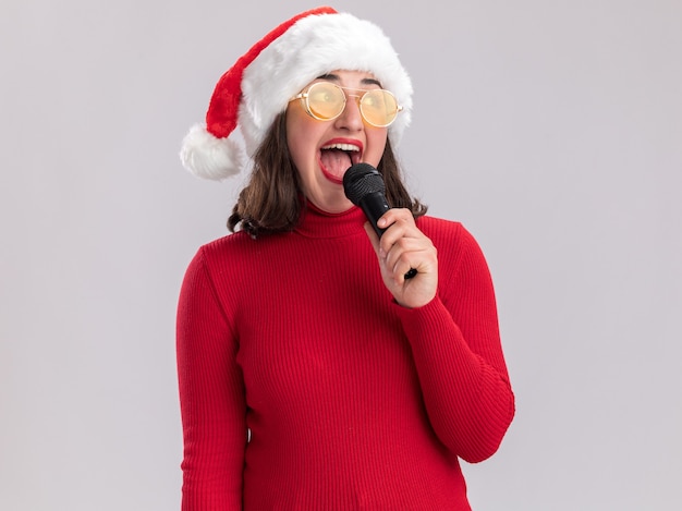 Szczęśliwa i wesoła młoda dziewczyna w czerwonym swetrze i santa hat w okularach trzymając mikrofon śpiewa piosenkę stojącą na białym tle