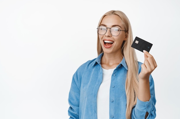 Szczęśliwa klientka banku w okularach pokazująca kartę kredytową gotową na zakupy stojącą na białym tle
