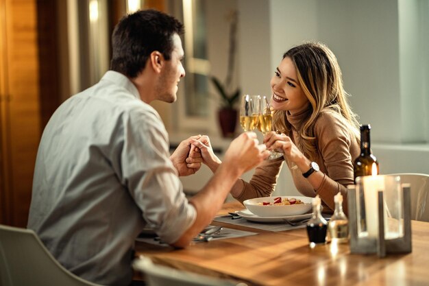 Szczęśliwa kobieta i jej chłopak trzymają się za ręce podczas opiekania szampanem podczas kolacji przy stole
