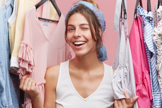 Szczęśliwa kobieta mrugając oczami stojąc w pobliżu regałów z ubraniami, bawiąc się i pozytywnie emocje po udanych zakupach