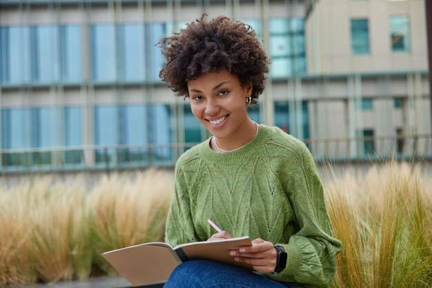 Szczęśliwa kobieta o kręconych włosach pracuje nad pisaniem eseju, zapisuje pomysły w notatniku, nosi swobodny zielony sweter, uśmiecha się pozytywnie do aparatu pozuje na zewnątrz na rozmytym tle, tworzy publikację tekstu