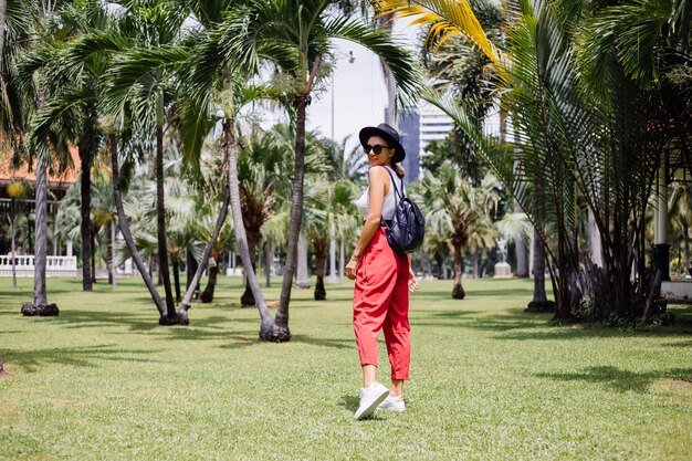 Szczęśliwa kobieta podróżuje po Bangkoku z plecakiem, ciesząc się pięknym słonecznym dniem w tropikalnym parku na zielonej trawie