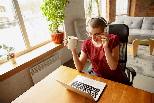 Szczęśliwa kobieta pracująca w domu podczas kwarantanny koronawirusa lub COVID-19, koncepcja zdalnego biura. Piękny łysy model, kierownik wykonujący zadania z laptopem, telefonem, ma konferencję online, spotkanie.