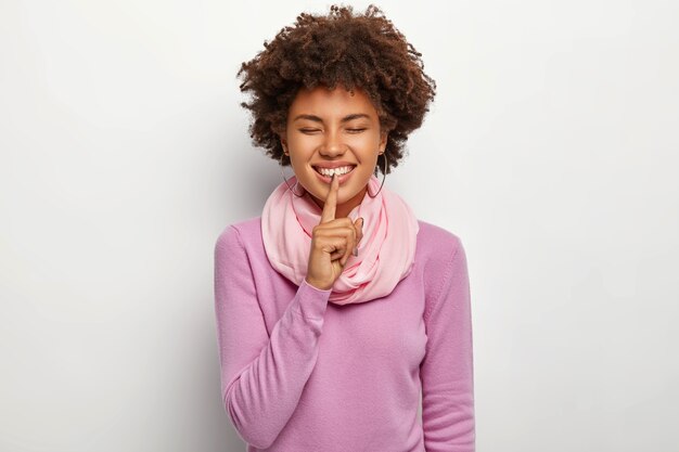 Szczęśliwa kobieta robi gest ciszy, trzyma palec wskazujący na ustach, cieszy się, że może podzielić się przyjemnym sekretem z najlepszą przyjaciółką, nosi fioletowy swobodny sweter