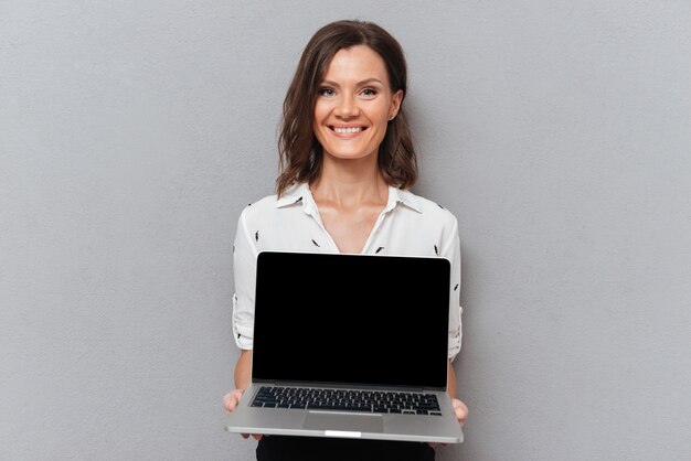 Szczęśliwa kobieta w biznesie odziewa pokazywać pustego laptopu ekran na popielatym