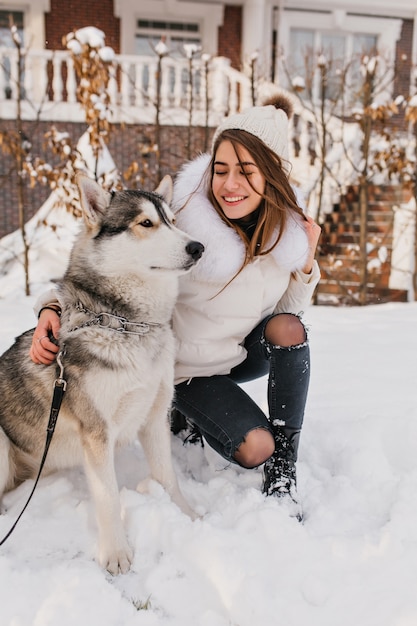 Szczęśliwa kobieta w czarnych dżinsach siedzi na śniegu po zabawnej grze z husky. Zewnątrz portret chilling europejskiej kobiety z psem w weekend grudnia.