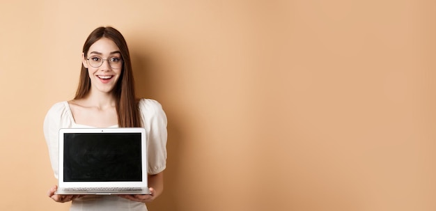 Bezpłatne zdjęcie szczęśliwa kobieta w okularach pokazująca ekran laptopa i uśmiechająca się demonstruje internetową ofertę promocyjną na stojąco