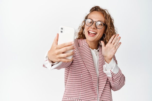 Szczęśliwa kobieta w okularach prowadząca rozmowę wideo, machająca do ekranu telefonu komórkowego i uśmiechnięta przyjaznym, białym tle