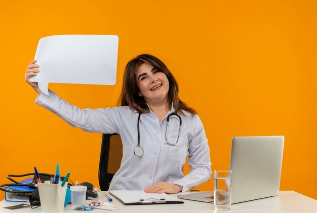 Szczęśliwa kobieta w średnim wieku ubrana w szlafrok medyczny i stetoskop siedzi przy biurku ze schowkiem na narzędzia medyczne i laptopem trzymając bańkę czatu na białym tle