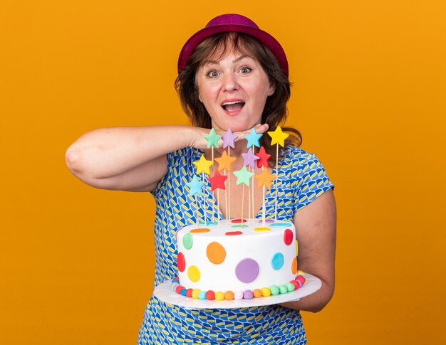 Szczęśliwa kobieta w średnim wieku w imprezowym kapeluszu trzymająca tort urodzinowy uśmiechnięta radośnie