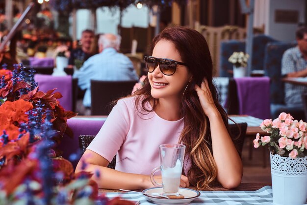 Szczęśliwa kobieta w średnim wieku z długimi brązowymi włosami, ubrana w różową sukienkę, siedząca ze szklanką cappuccino w kawiarni na świeżym powietrzu.