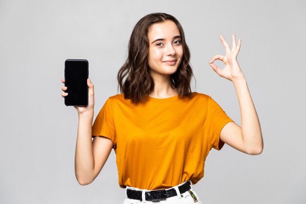 Szczęśliwa kobieta w ubranie pokazuje pusty ekran smartfona z w porządku gestem na szarej ścianie