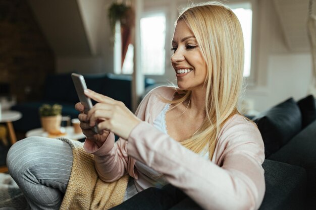 Szczęśliwa kobieta wysyła SMS-y na smartfonie, relaksując się w salonie