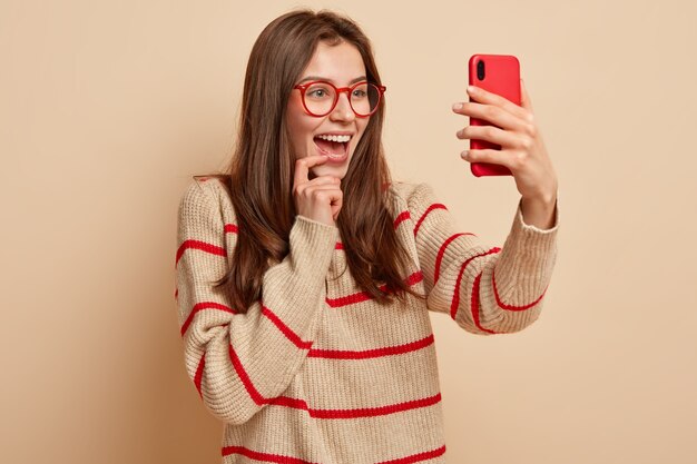 Szczęśliwa ładna dziewczyna robi śmieszne fotki, klika zdjęcie selfie na nowoczesnym telefonie komórkowym, publikuje posty w sieci społecznościowej, lubi się fotografować, nosi przezroczyste okulary, nosi swobodny sweter, izolowana