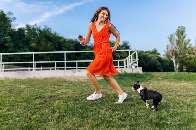 Szczęśliwa ładna kobieta w parku biegnie z psem boston terrier, uśmiechnięty pozytywny nastrój, modny letni styl, ubrana w pomarańczową sukienkę, bawi się ze zwierzakiem, dobra zabawa, kolorowe, aktywne wakacje weekendowe, trampki