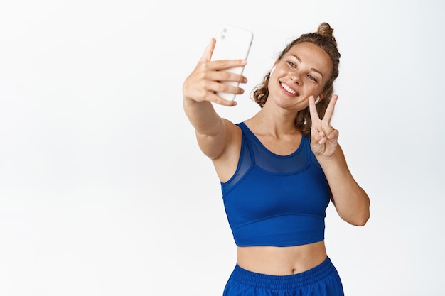 Szczęśliwa lekkoatletka biorąc selfie na telefon komórkowy, fotografując w słuchawkach i odzieży sportowej, białe tło